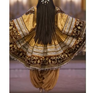 Può includere: Un tradizionale abito ricamato oro e marrone con maniche lunghe e gonna svasata. L'abito presenta motivi intricati, paillettes e un salwar dorato abbinato. La persona indossa un copricapo argentato e ha lunghi capelli scuri.