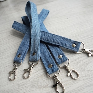 Könnte beinhalten: Ein Set aus vier Schlüsselbändern aus blauem Denim-Stoff, jeweils mit einem silberfarbenen, drehbaren Karabinerhaken, der mit einer silbernen Metallniete befestigt ist. Die Bänder bestehen aus strapazierfähigem Jeansstoff mit sichtbarer Köperbindung.