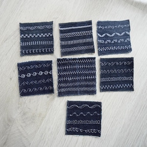 Op de afbeelding: Zeven vierkante lapjes spijkerstof gerangschikt in een groep. Elk donkerblauw lapje is voorzien van meerdere rijen witte decoratieve machinale borduursteken, waaronder golvende lijnen, lussen, zigzagpatronen en rechte lijnen op het oppervlak.