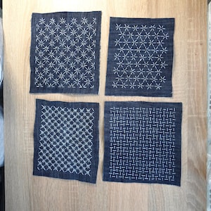 Può includere: Quattro pezzi di tessuto rettangolari blu scuro con motivi ricamati a mano in bianco. Ogni pezzo presenta un design geometrico distinto, inclusi motivi a forma di stella e schemi a griglia realizzati con punti filza dritti.