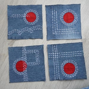 Könnte beinhalten: Vier quadratische Untersetzer aus Jeansstoff mit handgestickten weißen geometrischen Mustern und kreisförmigen roten Stoffapplikationen. Jeder Untersetzer zeigt eine einzigartige Anordnung aus weißen Strichlinien und einem roten Kreis auf blauem Denim.
