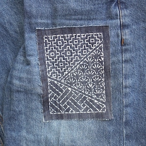 Könnte beinhalten: Eine blaue Jeans aus Denim mit einem rechteckigen, dunkelblauen Stoffaufnäher auf der Vorderseite. Der Aufnäher ist mit weißen, gestickten geometrischen Mustern verziert, darunter Kreuzformen, fächerartige Bögen und diagonale Linien.