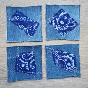 Könnte beinhalten: Vier quadratische Jeansstoff-Flicken, die jeweils mit einem mittig aufgenähten, blau-weißen Paisley-Stoffstück versehen sind, das mit sichtbaren Zickzackstichen befestigt wurde. Der Jeansstoff ist mittelblau und die Paisley-Stücke variieren in ihrer Form.