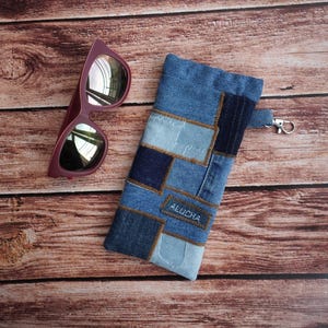 Pode incluir: Um estojo retangular de ganga com um design de retalhos em vários tons de azul, costuras castanhas e um pequeno fecho de mosquetão metálico. A palavra ALUCHA está bordada num remendo. Ao lado estão óculos de sol vermelho-escuro com lentes refletoras.