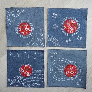 Könnte beinhalten: Vier quadratische Untersetzer aus Jeansstoff mit weißen handgestickten Mustern und einer runden Applikation aus rotem Blumenstoff in der Mitte. Die Stickereien zeigen geometrische Linien, kleine Kreuze und kreisförmige Motive.