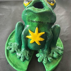 Può includere: Statuetta di rana verde fatta a mano con occhi gialli e una stella gialla sul petto, seduta su una foglia di ninfea verde. La statuetta ha il testo "@banshee_tears" su un lato.