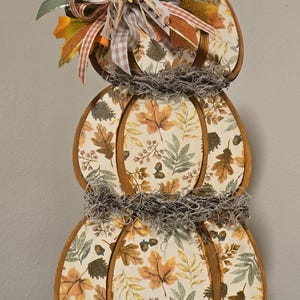 May include: A three-tiered pumpkin-shaped wooden decoration featuring a cream background with a pattern of orange, brown, and green autumn leaves and acorns. It is adorned with gray Spanish moss and a multi-layered ribbon bow with a small sunflower.