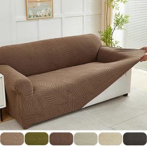 以下が含まれることがあります： 葉の模様が入った茶色のソファカバー。カバーはソファにかけられており、そのフィット感とデザインを示しています。カバーはニュートラルカラーで、さまざまなインテリアスタイルに適しています。いくつかのカラーオプションが画像の下部に表示されています。