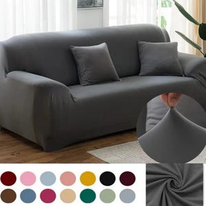 May include: A gray sofa cover with two matching throw pillows. The cover is stretched over the sofa, demonstrating its elasticity. A color palette of various shades is displayed below the sofa.