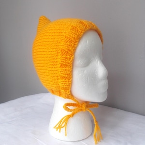 May include: A bright yellow knitted bonnet featuring a pointed top and a ribbed border along the face opening. The headwear includes two matching yellow yarn ties that are knotted together under the chin, each ending in a small decorative tassel.