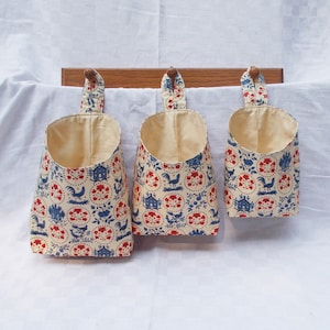 May include: Three hanging fabric storage baskets with a white background and a blue and red pattern of houses, flowers, and roosters.