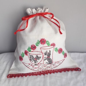 May include: White linen drawstring bag with red ribbon ties and red crocheted trim. The bag is embroidered with a design of two black silhouettes of dogs in cups, surrounded by red flowers and green leaves.