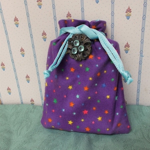 May include: A purple drawstring bag with a colorful star pattern. The bag is adorned with a decorative metal brooch with three light blue gems and a light blue ribbon drawstring. The bag is made of a soft, plush material.