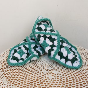 May include: Handmade crochet slippers in a granny square pattern. The slippers feature a combination of white and dark green squares, outlined with a matching green border. The slippers are displayed on a cream-colored, crocheted surface.