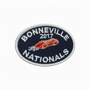 Peut inclure: Un écusson ovale avec les mots "BONNEVILLE NATIONALS" en blanc sur un fond bleu marine. L'année "2017" est au-dessus d'un dessin de voiture de course rouge. L'écusson a une bordure blanche cousue.