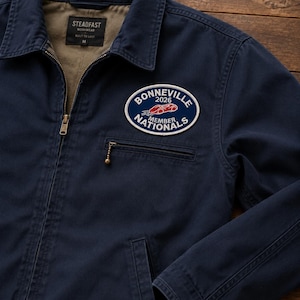 Può includere: Una giacca blu navy con cerniera, fodera interna color cuoio e colletto. Una toppa ovale sul petto mostra la scritta BONNEVILLE 2026 NATIONALS MEMBER con il grafico di un'auto da corsa rossa. L'etichetta interna riporta STEADFAST WORKWEAR.
