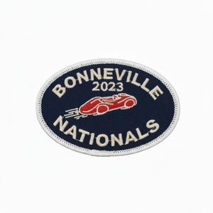 Peut inclure: Écusson ovale bleu marine avec l'inscription blanche "BONNEVILLE NATIONALS" et un dessin de voiture de course rouge. L'année "2023" est au-dessus de la voiture. Bordure blanche cousue.