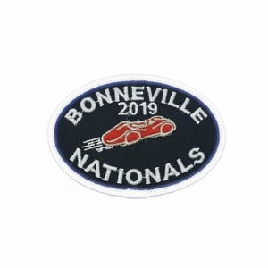 Peut inclure: Écusson ovale avec l'inscription "BONNEVILLE NATIONALS" en blanc sur fond bleu marine. Un dessin de voiture de course rouge est au centre, avec l'année "2019" au-dessus. L'écusson a une bordure blanche.