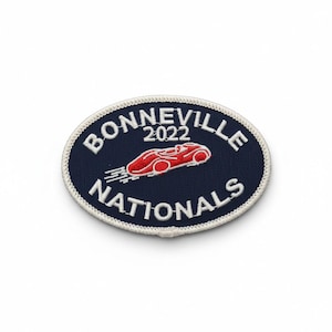 Puede incluir: Parche ovalado con las palabras "BONNEVILLE NATIONALS" en letras blancas sobre un fondo azul marino. El año "2022" está encima de un gráfico de coche de carreras rojo. El parche tiene un borde blanco.