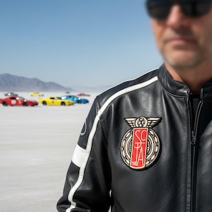 May include: Black leather jacket with white stripes and a patch featuring the letters "SC" and "TA". The jacket is worn by a person in front of colorful race cars on a salt flat.