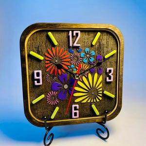 May include: A square wooden clock with rounded corners, featuring a dark stained finish. The clock face displays pink numerals at 12, 3, 6, and 9, with yellow markers for other hours. Colourful floral cutouts in orange, blue, purple, and yellow decorate the centre, accompanied by black hour and minute hands and a red second hand.