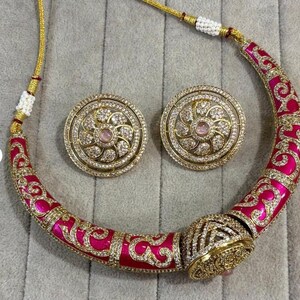 Puede incluir: Un conjunto de collar y pendientes dorados. El collar tiene un diseño curvo con un acabado de esmalte rosa brillante y detalles de filigrana dorada. Los pendientes son redondos, con una piedra rosa central y un halo de piedras claras.