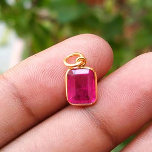 May include: A gold pendant featuring a rectangular, vivid pink gemstone. The gemstone is set within a gold frame, with a small loop at the top for a chain. The pendant is held in a hand against a blurred green backdrop.