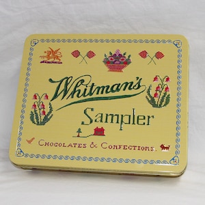 May include: A rectangular yellow metal Whitman's Sampler tin with a cross-stitch pattern design. The lid features the text Whitman's Sampler and Chocolates & Confections, surrounded by small illustrations of flowers, a house, a bird, and a dog.