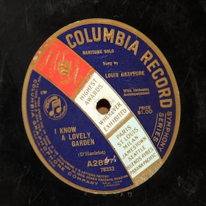 May include: A vintage Columbia Record 78 RPM record with a gold label. The label features a blue and gold design with the text "Columbia Record Symphony Series" and "I Know A Lovely Garden (D'Hardelot)". The record is by Louis Graveure and is from the A28974 series.