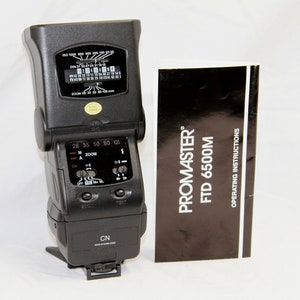 May include: A black Promaster FTD 6500M flash unit with a digital display and zoom controls. The flash unit is accompanied by a black and white instruction manual.