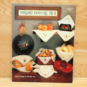May include: A book titled "Bread Covers IV" with charted designs by Harriette Tew. The book features a variety of bread covers with different designs, including a pineapple, a basket of fruit, and a loaf of bread. The book is on a wooden surface.