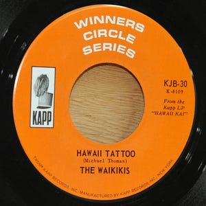 May include: A vintage vinyl record with an orange label featuring the text "WINNERS CIRCLE SERIES." The label includes the song title "HAWAII TATTOO" by The Waikikis, and the Kapp Records logo. The record is black.