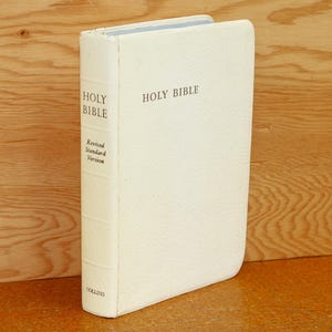 May include: Cream-colored Holy Bible book with the words "Holy Bible" printed on the cover and spine. The spine also reads "Revised Standard Version". The book is standing upright on a wooden surface.