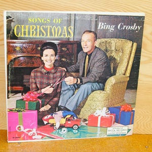May include: Vintage album cover for "Songs of Christmas" by Bing Crosby. The cover features a man and woman, surrounded by wrapped gifts and toy trucks. The title is in gold lettering.