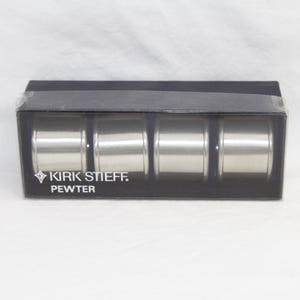 May include: A set of four pewter napkin rings by Kirk Stiff in a clear plastic box. The napkin rings are cylindrical and have a brushed metal finish. The box has a dark blue base and the brand name is printed in white.