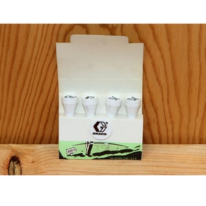 May include: A pack of four white golf tees with a black Graco logo and the text "The Action Line, U.S.A." on the bottom. The tees are in a white cardboard box with a green and white illustration of a golfer.