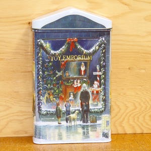 May include: A rectangular tin with a white roof and a festive illustration. The image depicts a snowy scene outside a shop called "TOY EMPORIUM", with figures, a dog, and a Christmas tree.