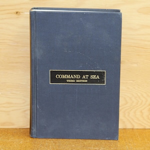 May include: A navy blue book titled "COMMAND AT SEA" with "THIRD EDITION" below. The book has a rectangular gold label with black text. The book is standing upright against a light brown wooden background.