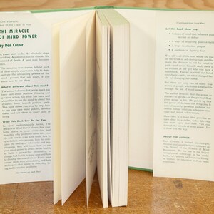 Op de afbeelding: Een open boek getiteld "The Miracle of Mind Power" door Dan Custer. Het boek heeft een groene rug en witte pagina's. Tekst op de linkerpagina bespreekt de inhoud van het boek, terwijl de rechterpagina opsomt wat het boek de lezer zal laten zien.