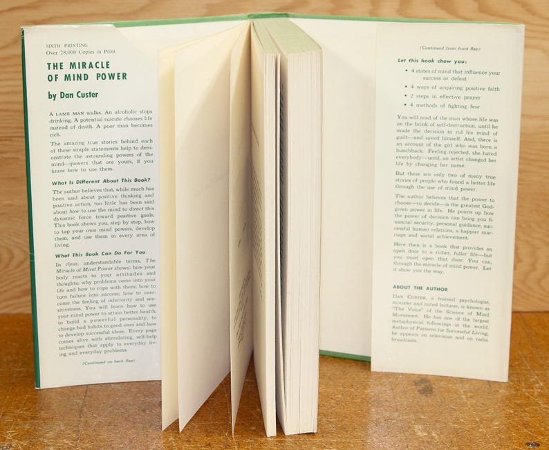 Op de afbeelding: Een open boek getiteld "The Miracle of Mind Power" door Dan Custer. Het boek heeft een groene rug en witte pagina's. Tekst op de linkerpagina bespreekt de inhoud van het boek, terwijl de rechterpagina opsomt wat het boek de lezer zal laten zien.