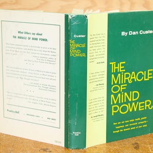 Op de afbeelding: Een open boek getiteld "The Miracle of Mind Power" van Dan Custer. De kaft is groen met gele tekst. De rug van het boek toont de titel en auteur. De linkerpagina bevat tekst over het boek. Het boek ligt op een houten oppervlak.