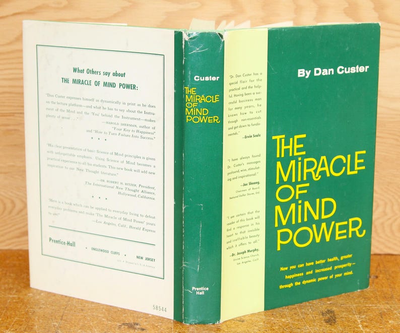 Op de afbeelding: Een open boek getiteld "The Miracle of Mind Power" van Dan Custer. De kaft is groen met gele tekst. De rug van het boek toont de titel en auteur. De linkerpagina bevat tekst over het boek. Het boek ligt op een houten oppervlak.