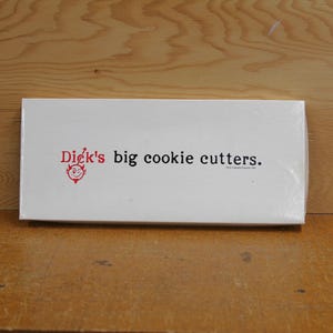 May include: A white rectangular box with the text "Dick's big cookie cutters." in black and red. A small red devil face logo is on the left. The box is on a wooden surface.