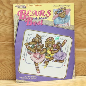 May include: A purple and white cross stitch pattern booklet featuring five designs of bears. The cover features a bear wearing a blue dress and a pink hat. The title of the booklet is "Bears at their Best".