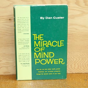 Op de afbeelding: Een hardcover boek getiteld "The Miracle of Mind Power" door Dan Custer. Het boek heeft een groene kaft met gele letters. De rug van het boek is lichtgroen met tekst. Het boek ligt op een houten oppervlak.