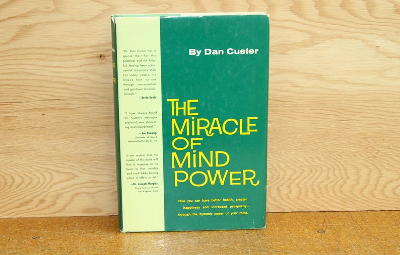 Op de afbeelding: Een hardcover boek getiteld "The Miracle of Mind Power" door Dan Custer. Het boek heeft een groene kaft met gele letters. De rug van het boek is lichtgroen met tekst. Het boek ligt op een houten oppervlak.