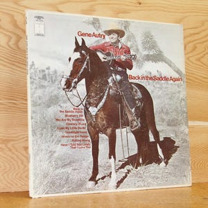 May include: Vintage vinyl record album cover featuring Gene Autry on horseback, playing a guitar. The album is titled "Back in the Saddle Again." The cover art is in sepia tones with red text. The album's tracklist is also visible.