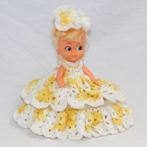 May include: A small doll wearing a white and yellow crocheted dress and hat. The dress has multiple layers and the hat is a flower shape.