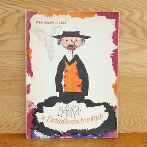 May include: A rectangular book titled Bajka o Dziadkowych wotach by Władysław Dulęba. The cover features a stylized illustration of a person wearing a black hat and orange vest, set against a white background with pink and orange watercolor-style borders.
