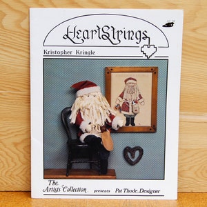 May include: A cross-stitch pattern featuring a Santa Claus figure with a red and white suit, white beard, and a bag of gifts. The pattern is titled "Kristopher Kringle" and is presented by "The Artis Collection" with Pat Thod as the designer.